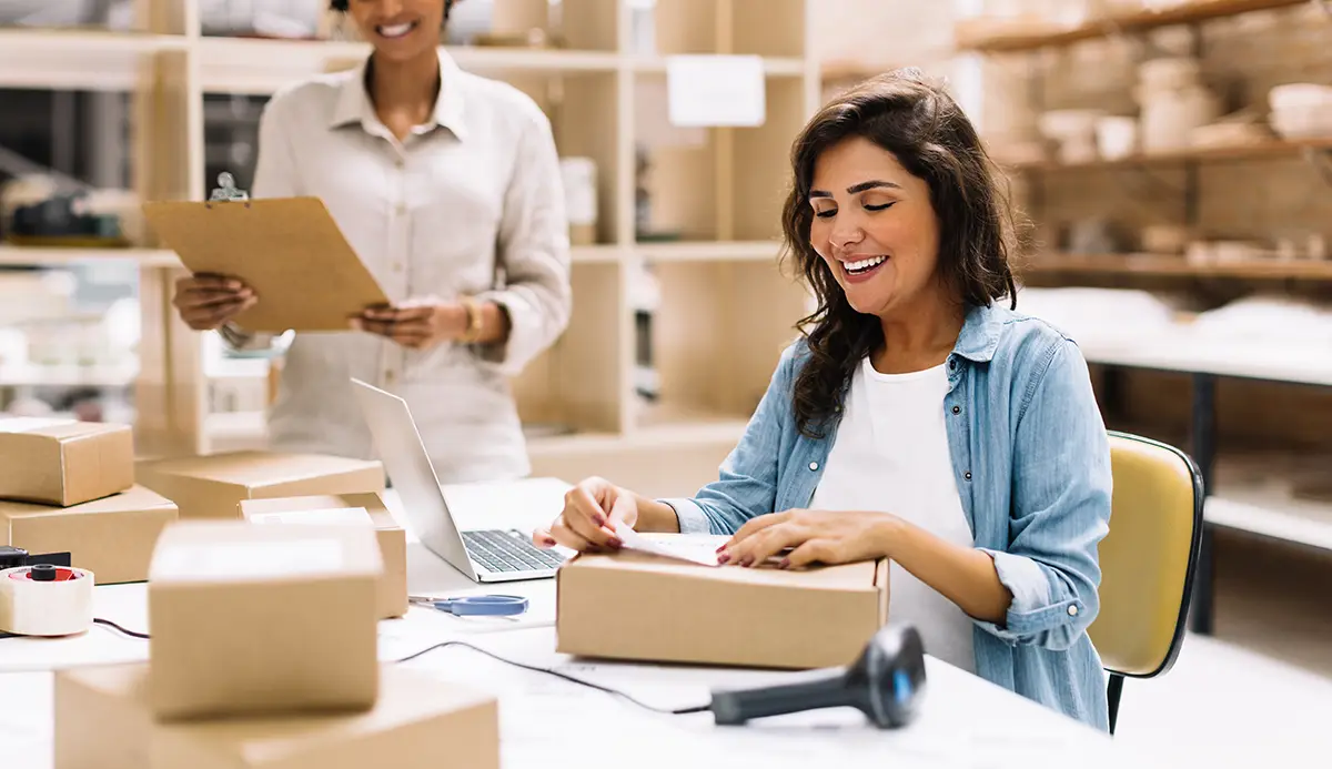 Happy female warehouse worker using best free multi-carrier shipping software Descartes XPS Ship to print a shipping label and apply it to a parcel as a co-worker looks on