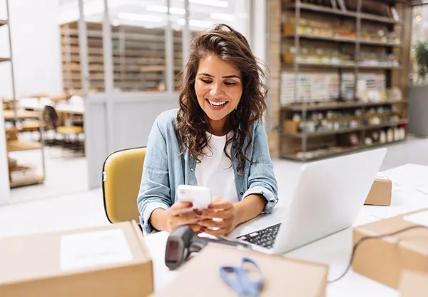 A happy ecommerce shipper in a warehouse with her laptop computer and smartphone just received a payout from a shipping insurance claim